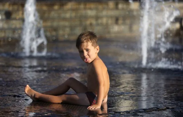 Мальчик играет в струях фонтана в жаркий летний день и показывает язык фотографу
