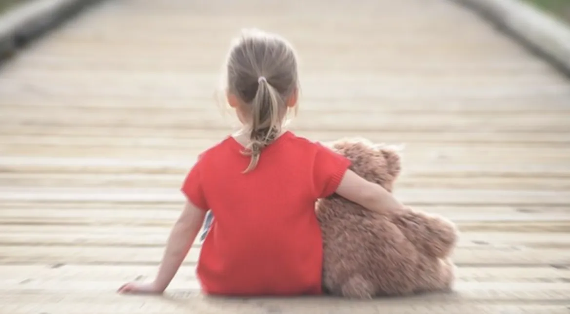 Девочка на деревянном настиле сидит спиной к зрителю с медвежонком под рукой